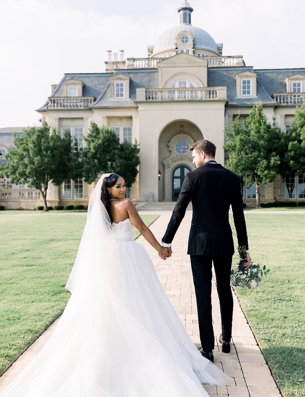 The bride and groom walk away holding hands down a path toward the grand French chateau-style wedding venue, showcasing the bride's long tulle train.