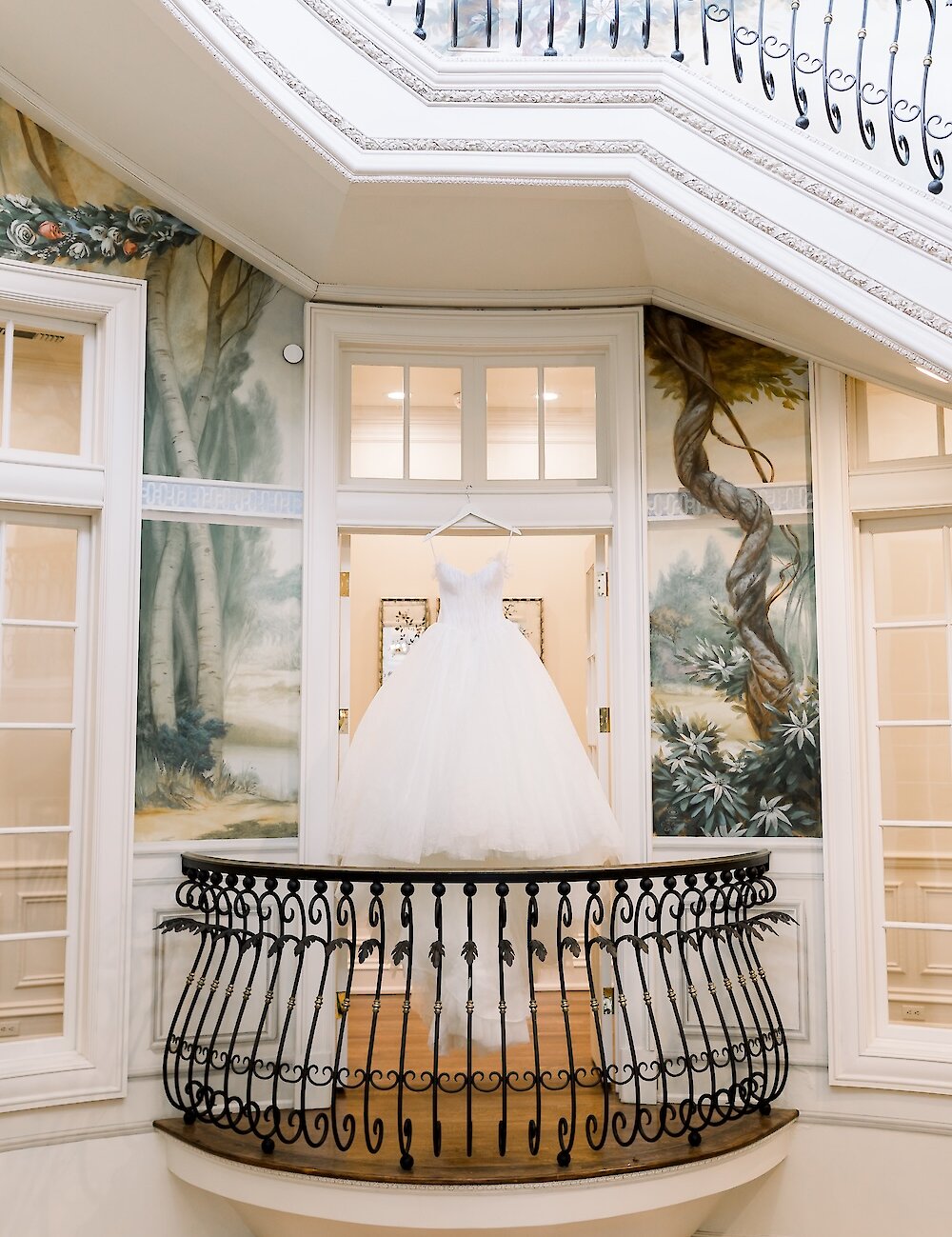 The bride's voluminous white ballgown hangs in an ornate doorway above a small, decorative metal balcony inside a luxurious venue with painted murals.