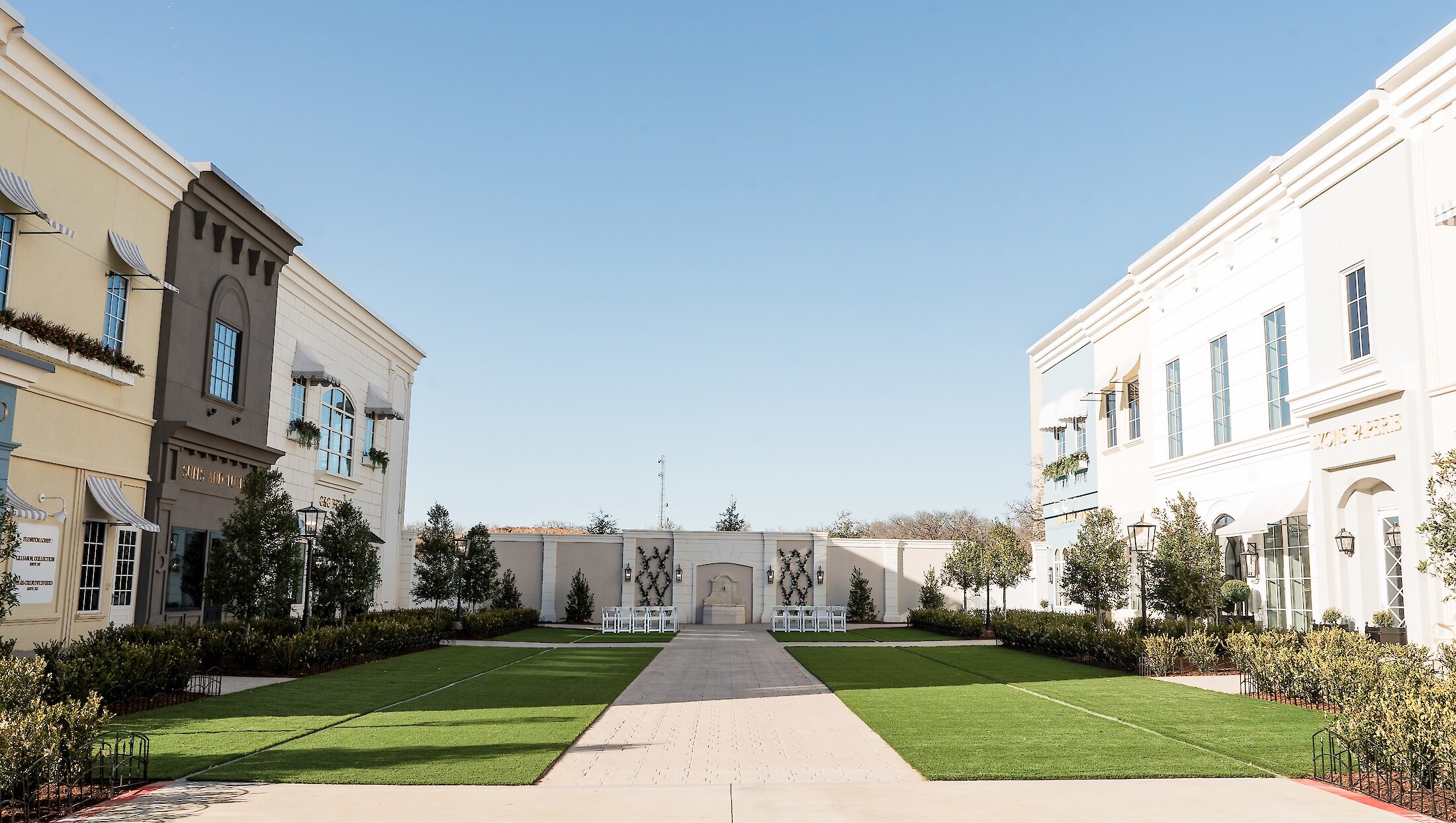 A wide shot of a European-style outdoor courtyard with buildings on both sides, a paved central aisle, green turf, and a decorated ceremony wall at the far end.