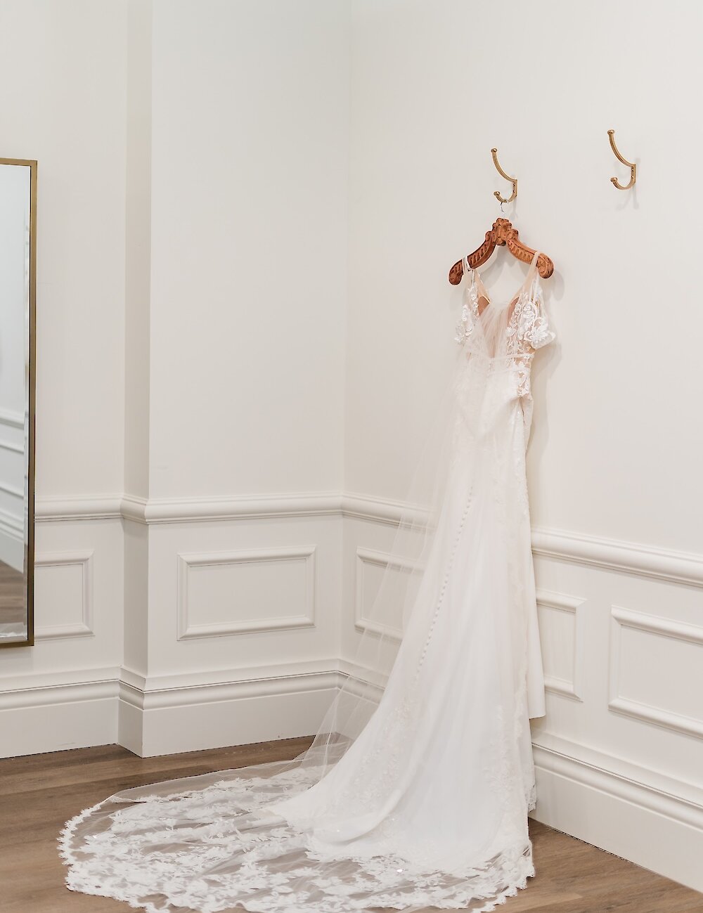 A white lace wedding dress with a long train hangs on a wooden hanger on a white wall with wainscoting and gold hooks in a bridal suite.