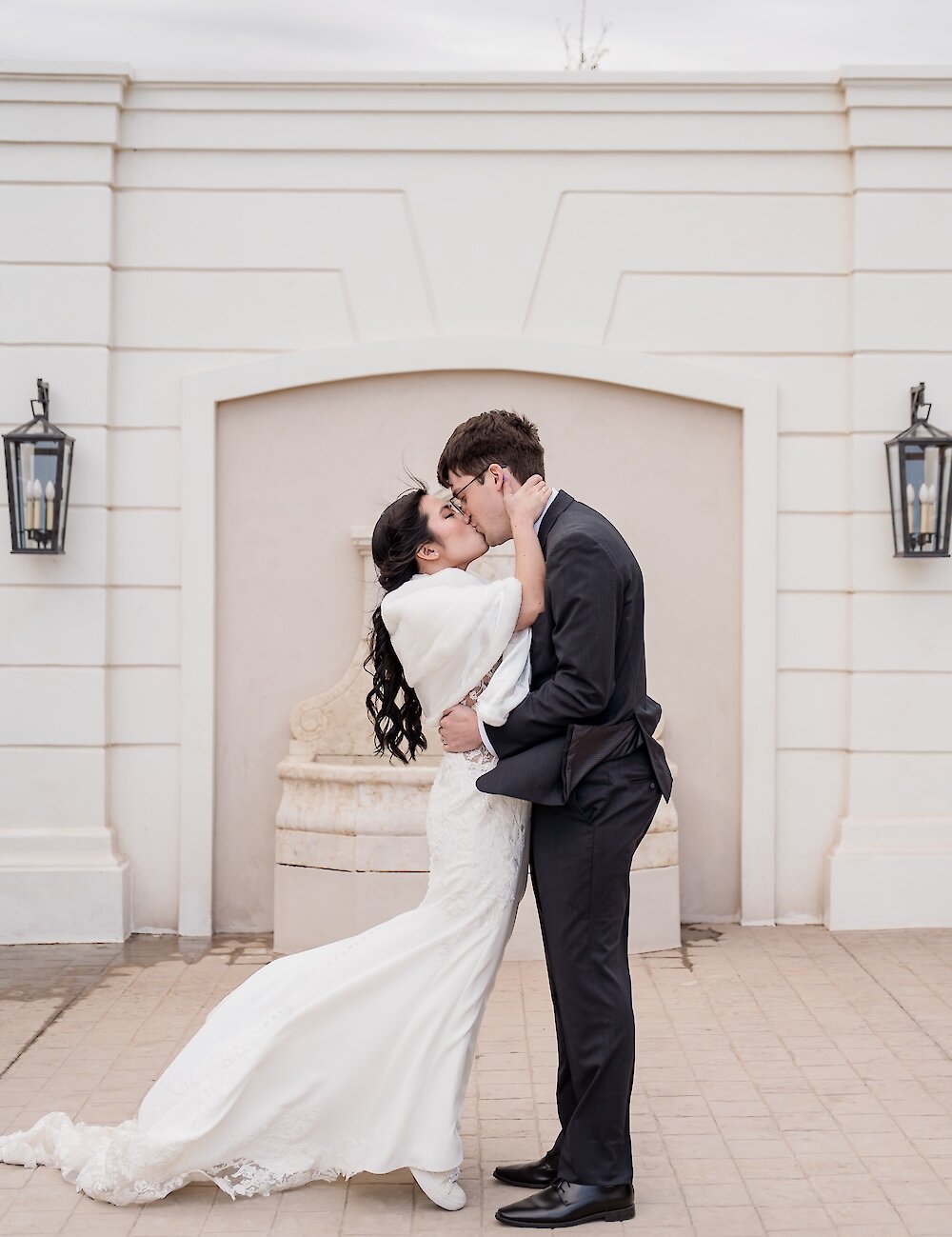 The bride in a white fur wrap and lace gown kisses the groom in a dark suit, standing on a patio in front of a white architectural wall with lanterns.