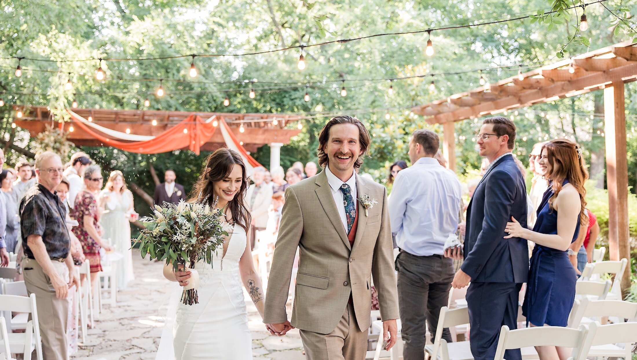 A joyful bride and groom walk hand-in-hand down the outdoor aisle, surrounded by smiling guests, under string lights and wooden pergolas.