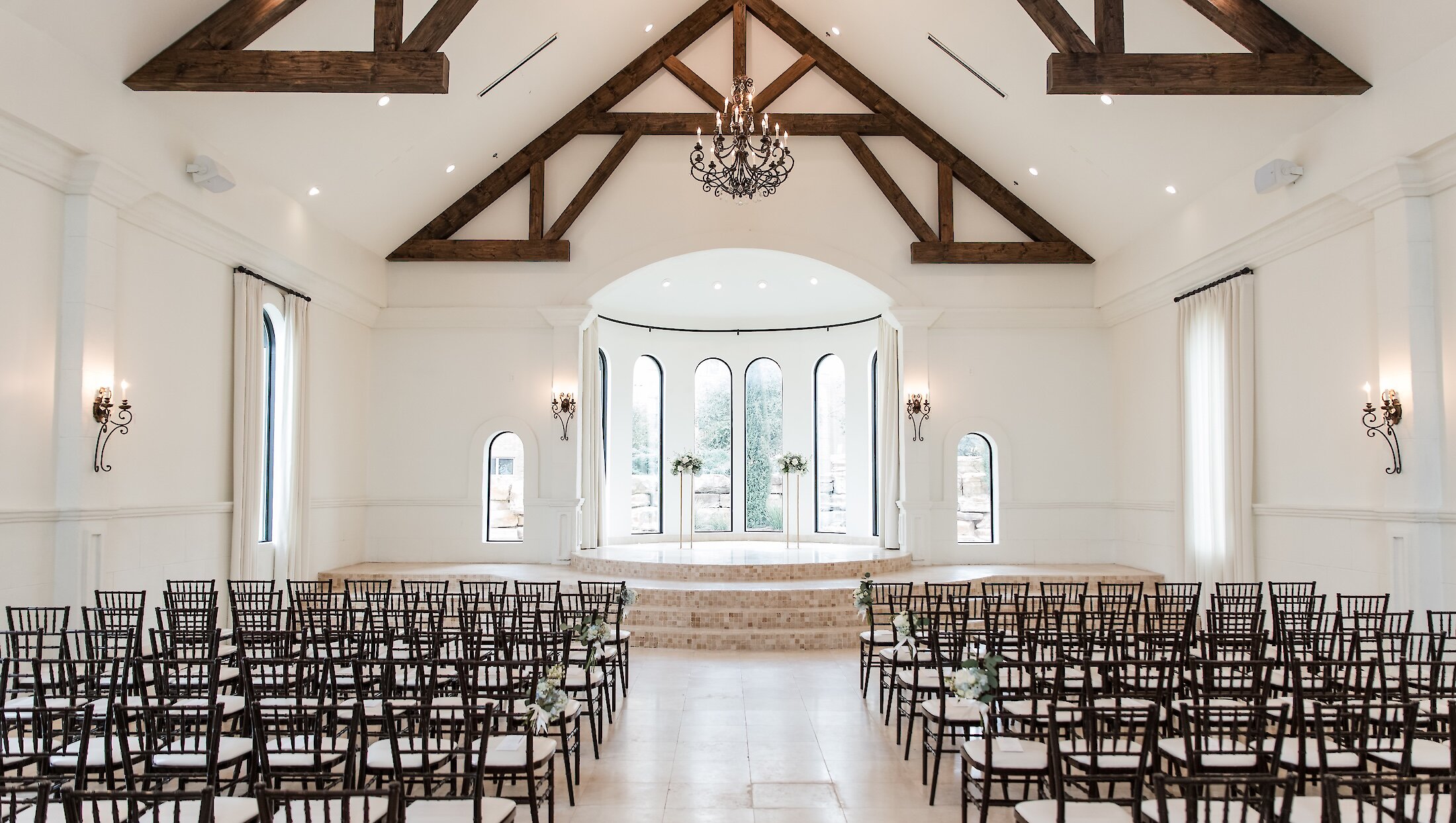 The elegant interior of a bright white chapel featuring dark wooden ceiling trusses, a tiered altar, and rows of dark wood chiavari chairs.