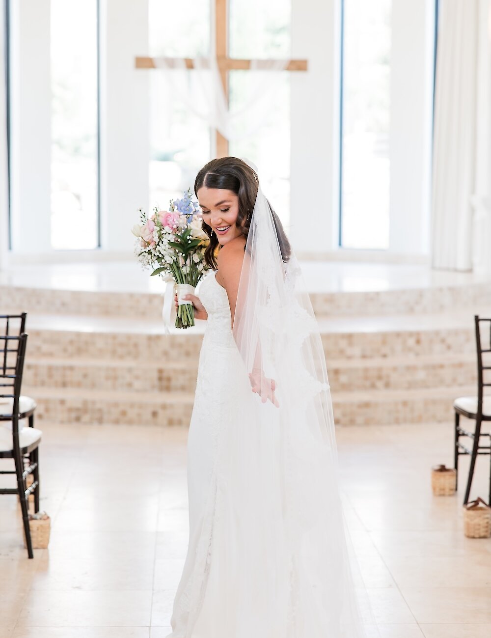 A smiling bride holds a colorful pastel bouquet and looks over her shoulder while standing in the aisle of a bright white chapel with a wooden cross altar.