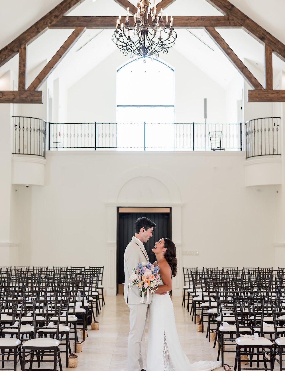 The bride and groom smile and embrace in the chapel aisle, with the groom in a tan suit and the bride holding a pastel bouquet, beneath a large chandelier.