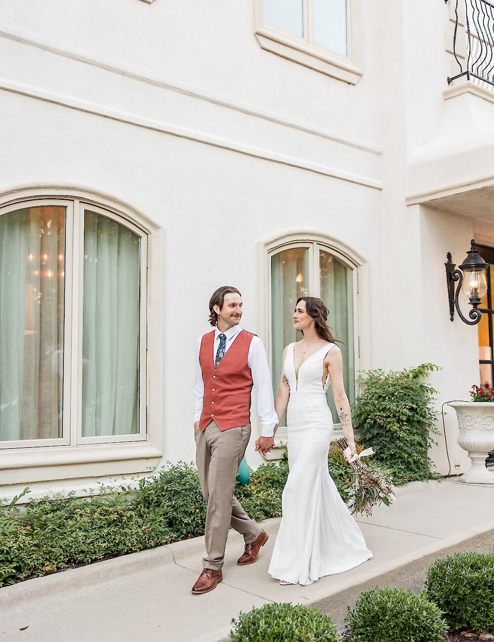 The bride and groom smile at each other while walking outside the venue, with the groom wearing a rust-colored vest and the bride a low-cut white gown.