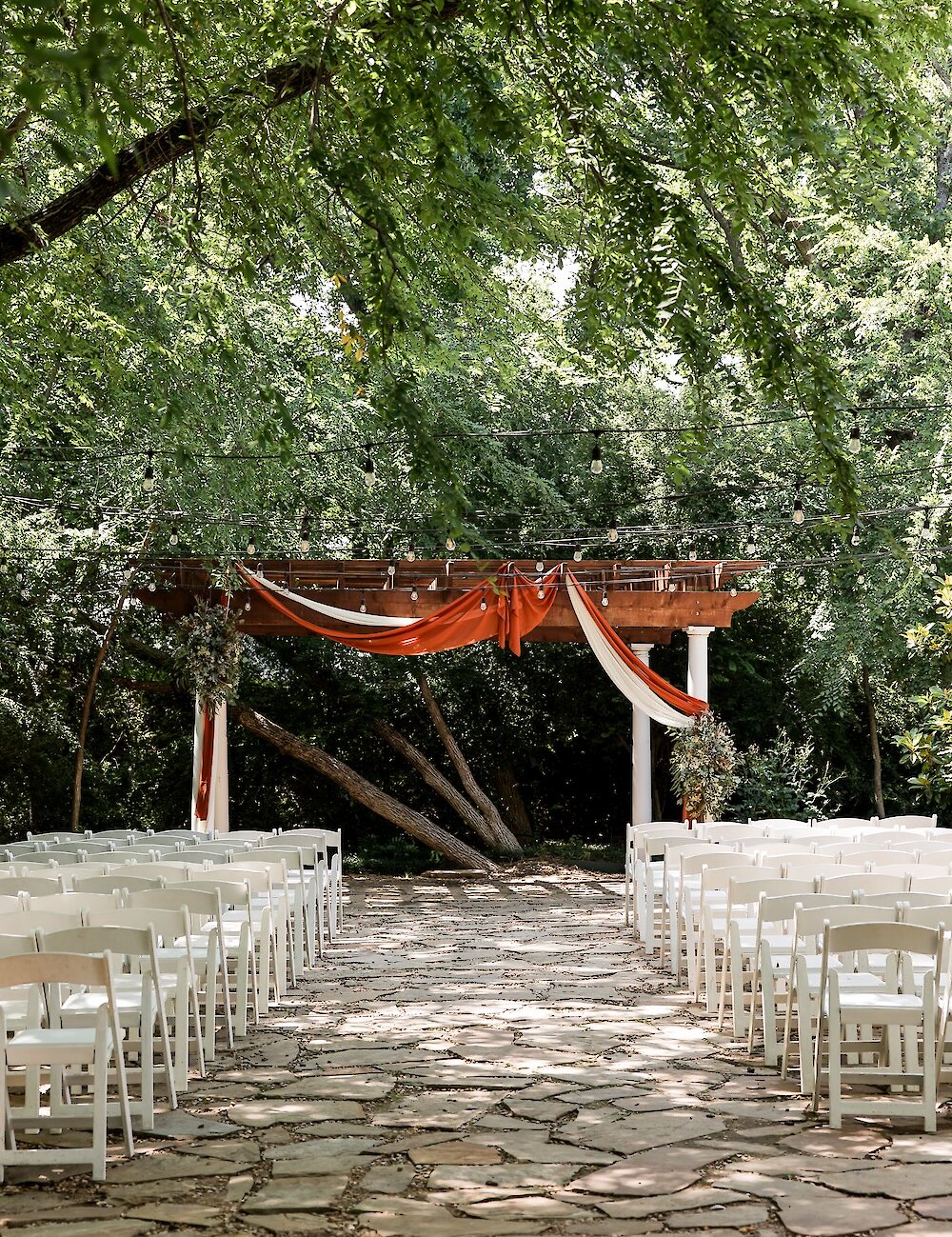 An outdoor wedding ceremony setup featuring a flagstone aisle, white folding chairs, and a wooden pergola draped with white and rust-colored fabric under a canopy of trees.