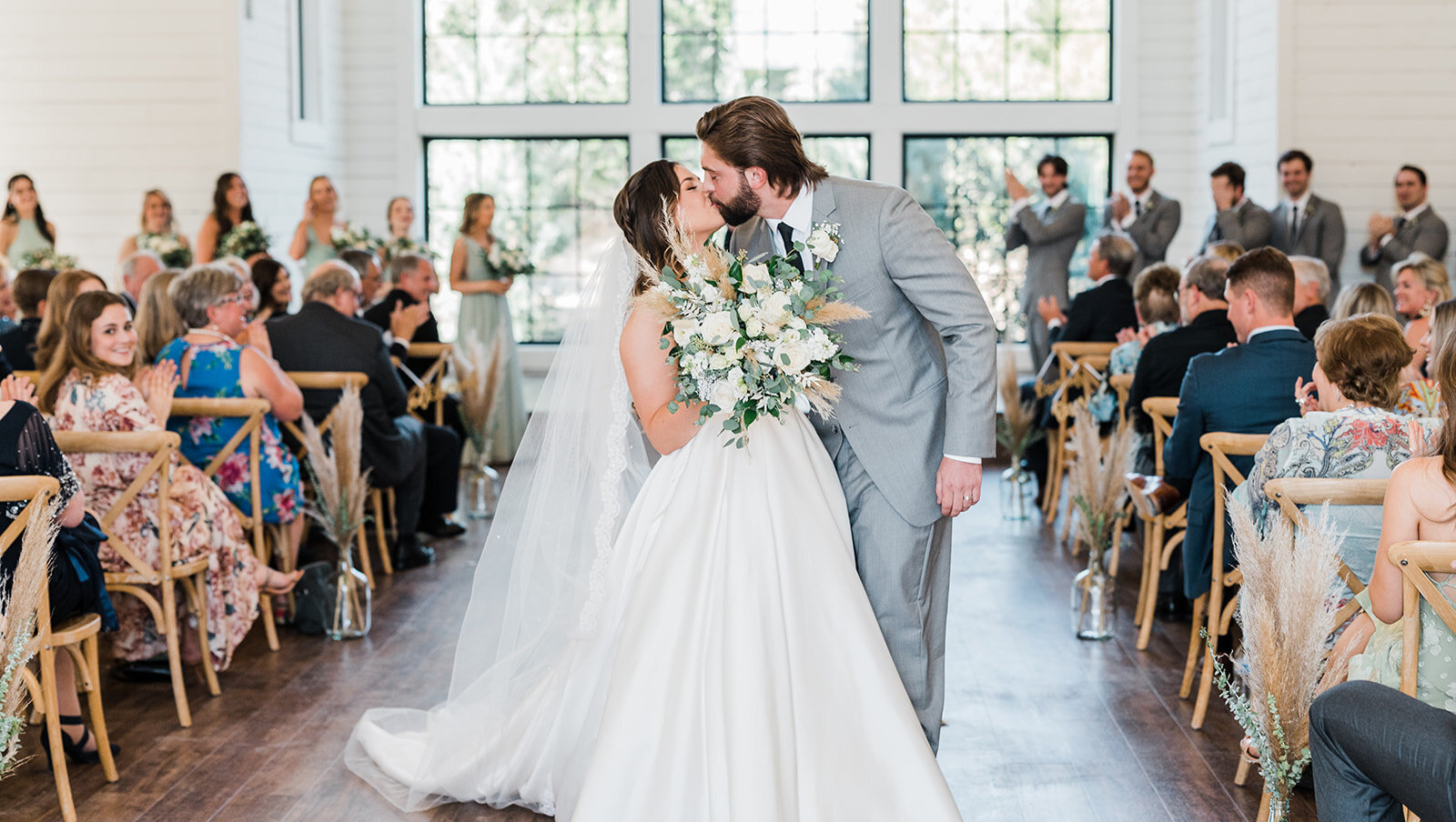 The bride and groom share a joyful first kiss in the aisle after the ceremony, surrounded by clapping guests in a bright white barn venue with large windows.