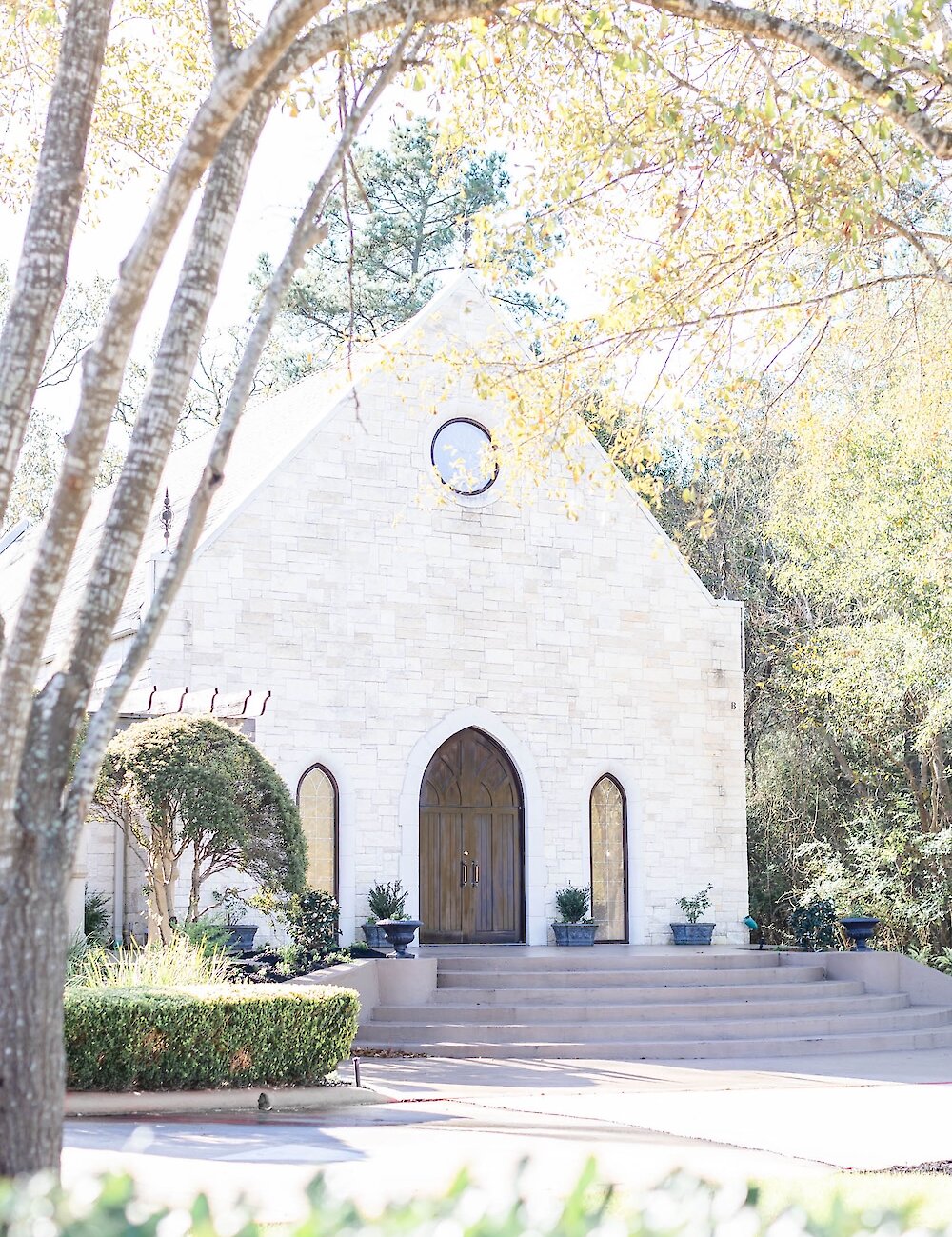 The picturesque exterior of a white stone chapel featuring a wooden arched door and steps, framed by green trees and foliage in the sunlight.