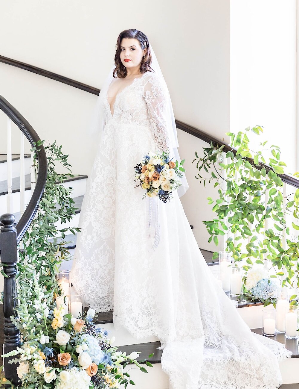 The bride in a long-sleeve lace gown stands on a dark wood spiral staircase adorned with greenery, candles, and blue and yellow floral arrangements.