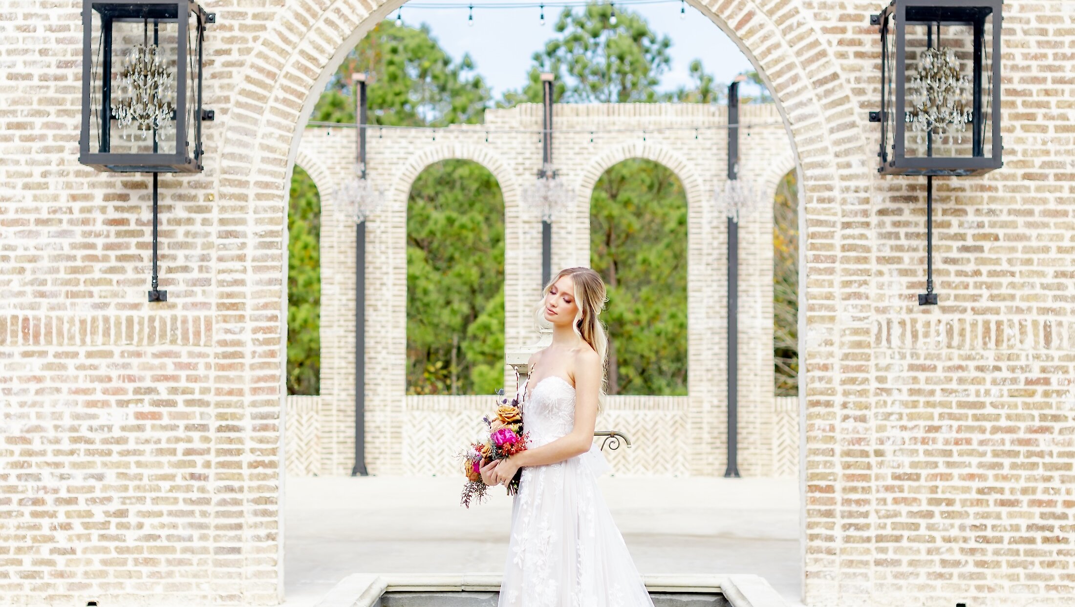 The bride in a strapless lace gown holds a colorful bouquet and stands framed by a large arched opening in a light brick wall structure.