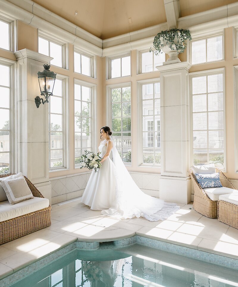 The beautiful bride in a white gown stands in a sunlit interior room holding a white bouquet, next to a small pool and wicker lounge furniture.