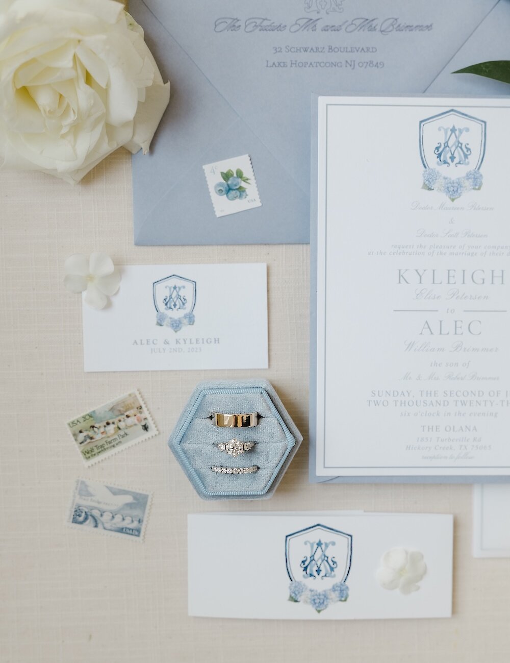 A wedding flat lay featuring the blue and white invitation suite with a monogram crest, the diamond engagement ring and bands in a blue velvet box, and white rose accents.
