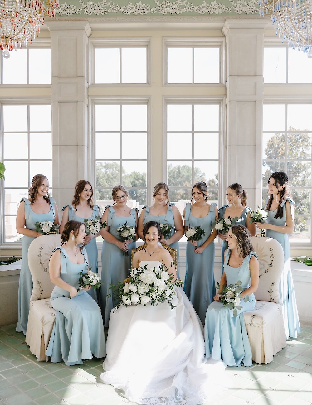 The bride sits holding a large white bouquet, surrounded by her nine bridesmaids in matching dusty blue dresses, posing in a sunlit room with tall arched windows and a mint green floor.