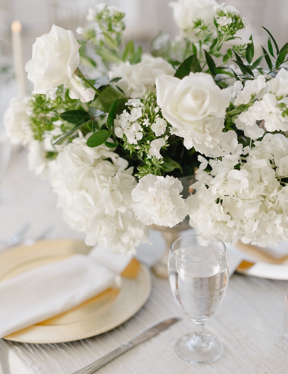 Close-up of a wedding reception place setting featuring a white floral centerpiece, a gold charger plate, a white napkin, and a stemmed water glass.