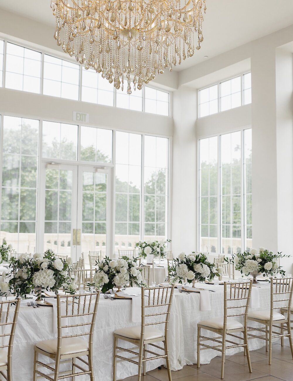An elegant reception hall with high windows, a crystal chandelier, and long tables set with white linens, gold chiavari chairs, and white floral centerpieces.