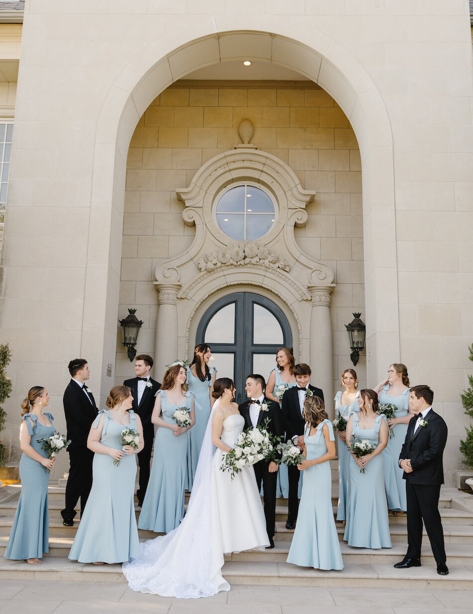 The bride and groom pose with their wedding party in blue dresses and black tuxedos on the sweeping stone steps of a grand mansion venue entrance.