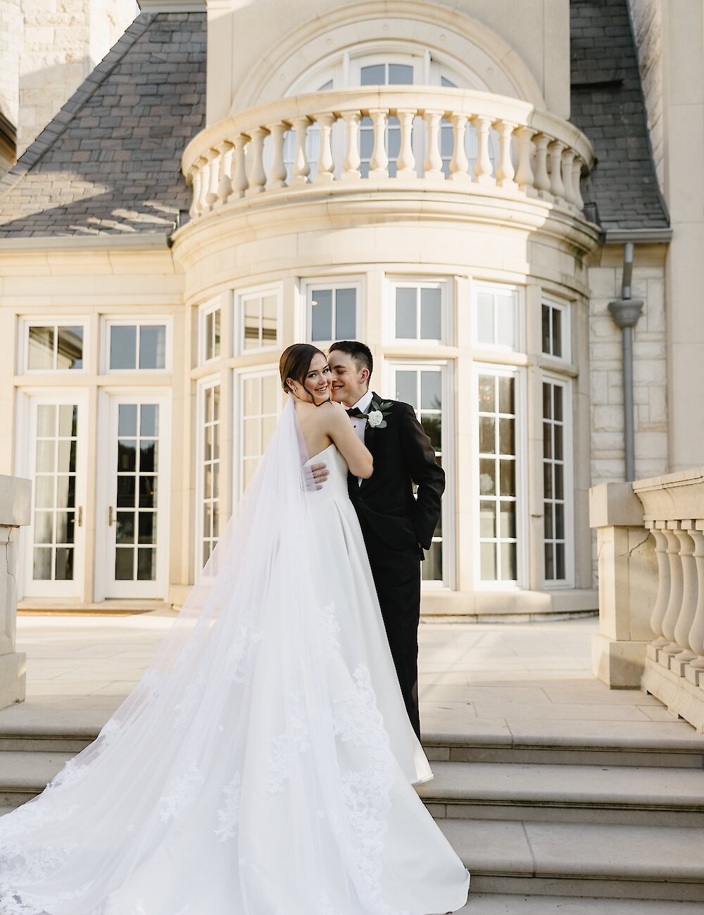 The bride smiles over her shoulder while embracing the groom on the stone steps outside the mansion's curved balcony and French doors.