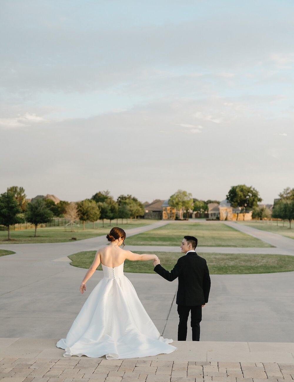 The bride and groom are walking away and holding hands down a wide stone staircase, leading to a sprawling manicured lawn and residential background.