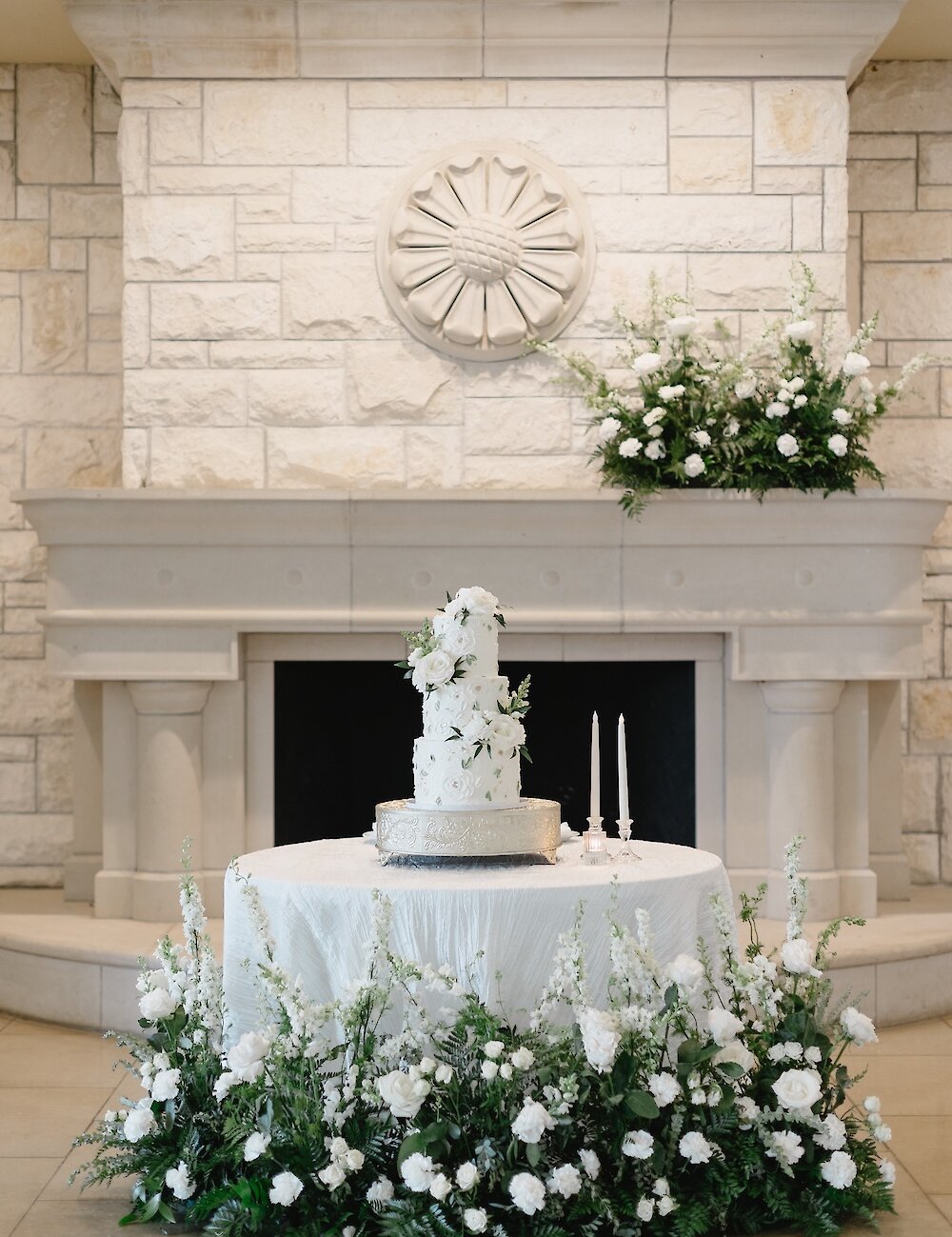 A tiered white wedding cake sits on a small round table in front of a white stone fireplace with a circular medallion and abundant white and green floral arrangements.