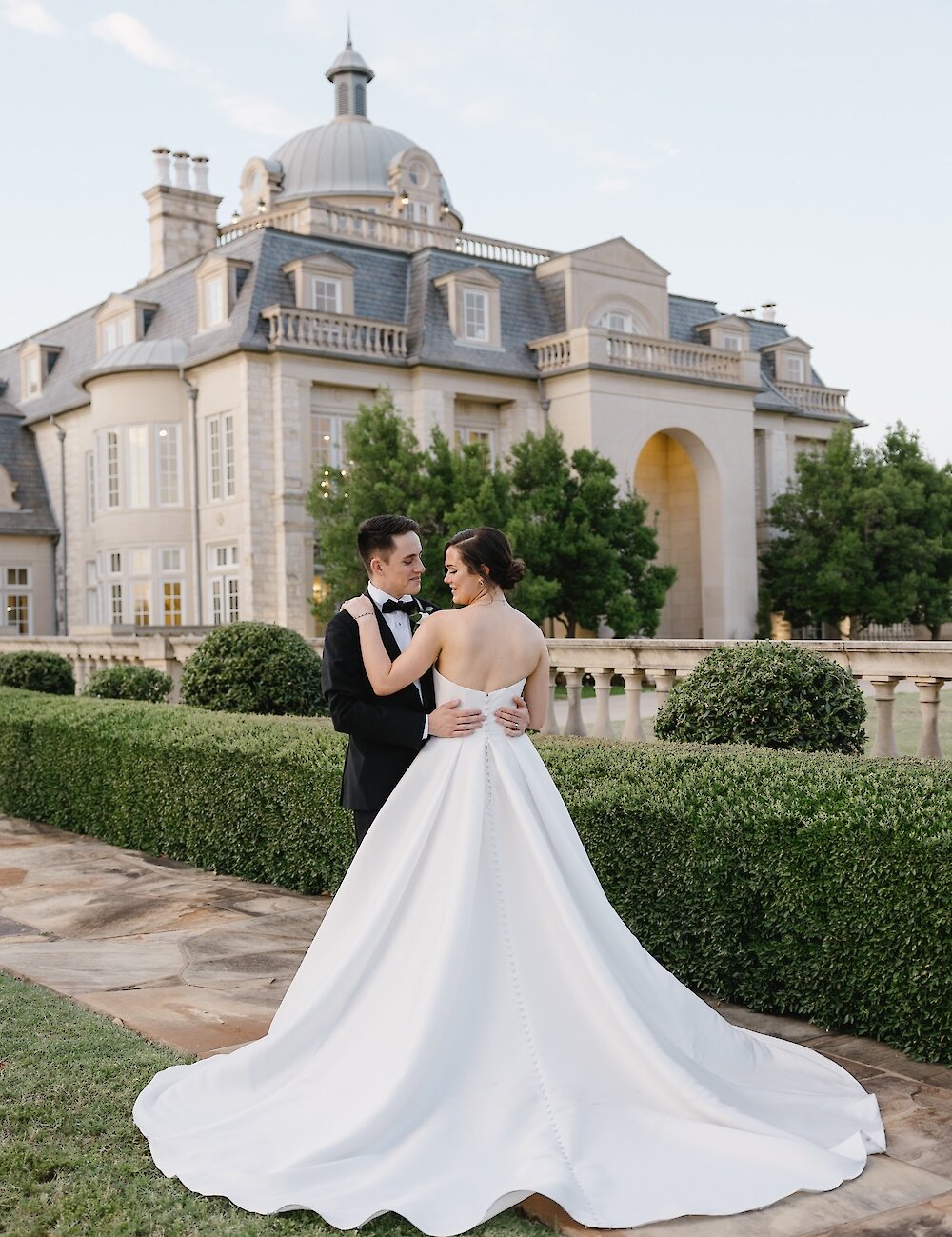 The bride and groom hug and gaze at each other during an evening portrait, with the bride's satin gown train spread out on the stone path in front of a French chateau mansion.