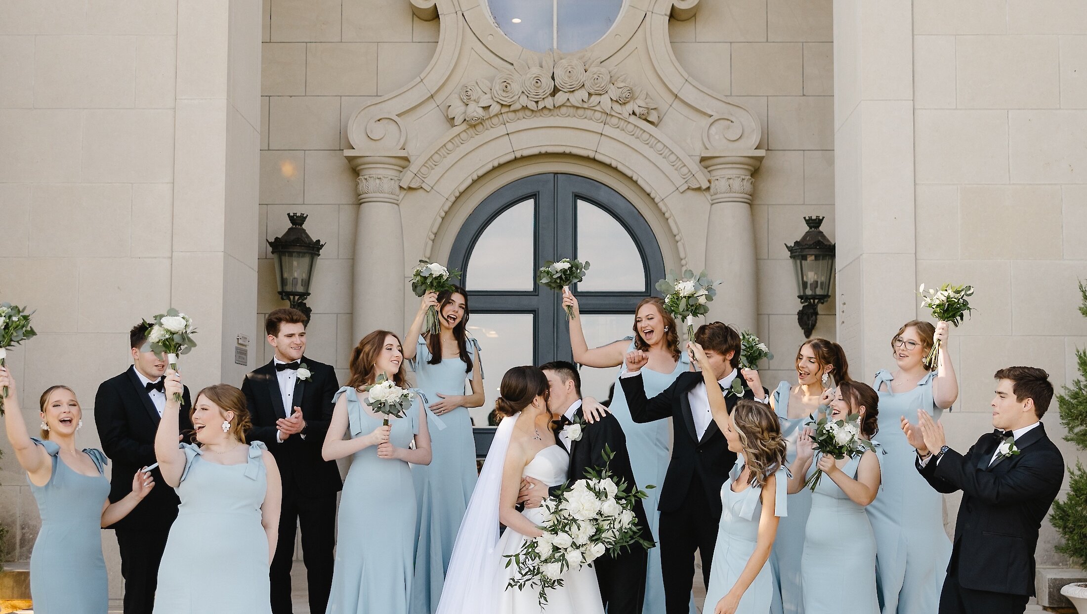 The bride and groom kiss on the steps of a grand stone mansion entrance, surrounded by their cheering wedding party in black tuxedos and light blue dresses.