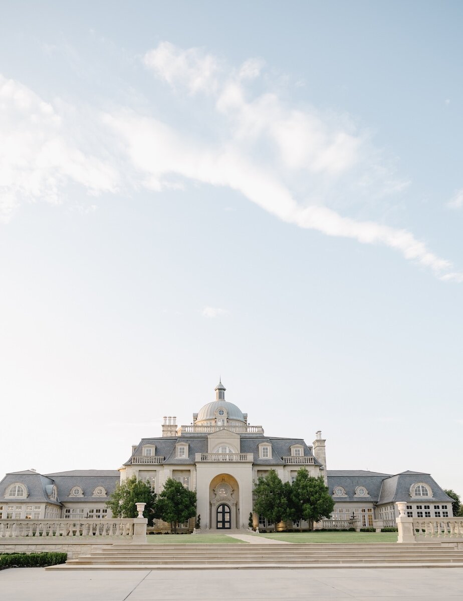 The magnificent exterior of a French chateau-style wedding mansion featuring a central dome, grand stone steps, and symmetrically placed trees on the lawn.
