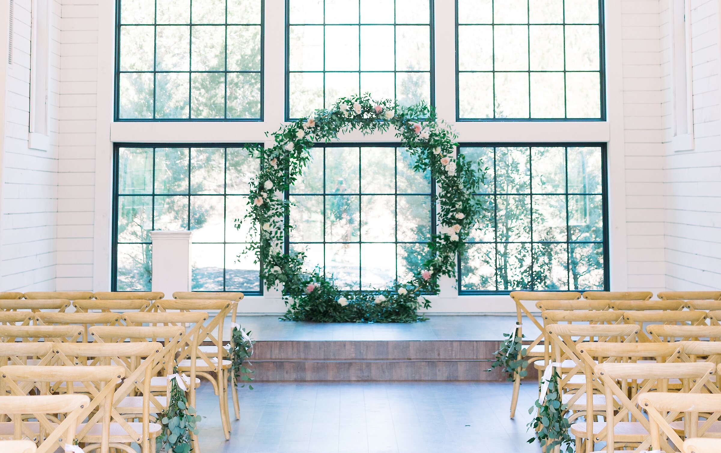 The interior of a white barn-style chapel featuring wooden cross-back chairs, a large chandelier, and a circular floral wreath altar in front of grid windows.