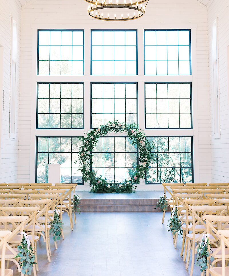 The interior of a white barn-style chapel featuring wooden cross-back chairs, a large chandelier, and a circular floral wreath altar in front of grid windows.