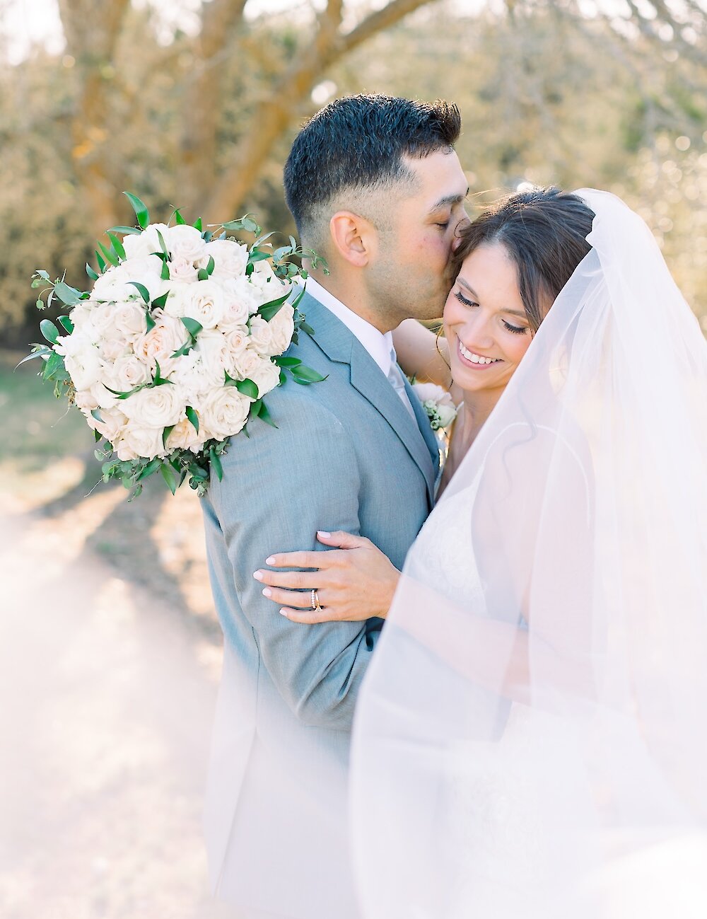The groom in a gray suit kisses the smiling bride on the forehead for a romantic outdoor portrait, with her veil and large white bouquet visible.