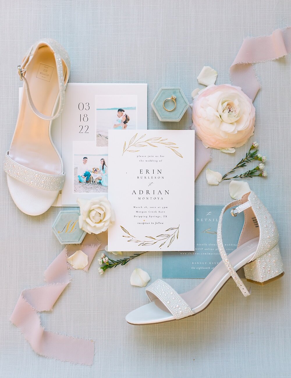 A wedding flat lay featuring white jeweled bridal shoes, gold-foiled invitations, the engagement ring in a velvet box, and white and blush floral accents.