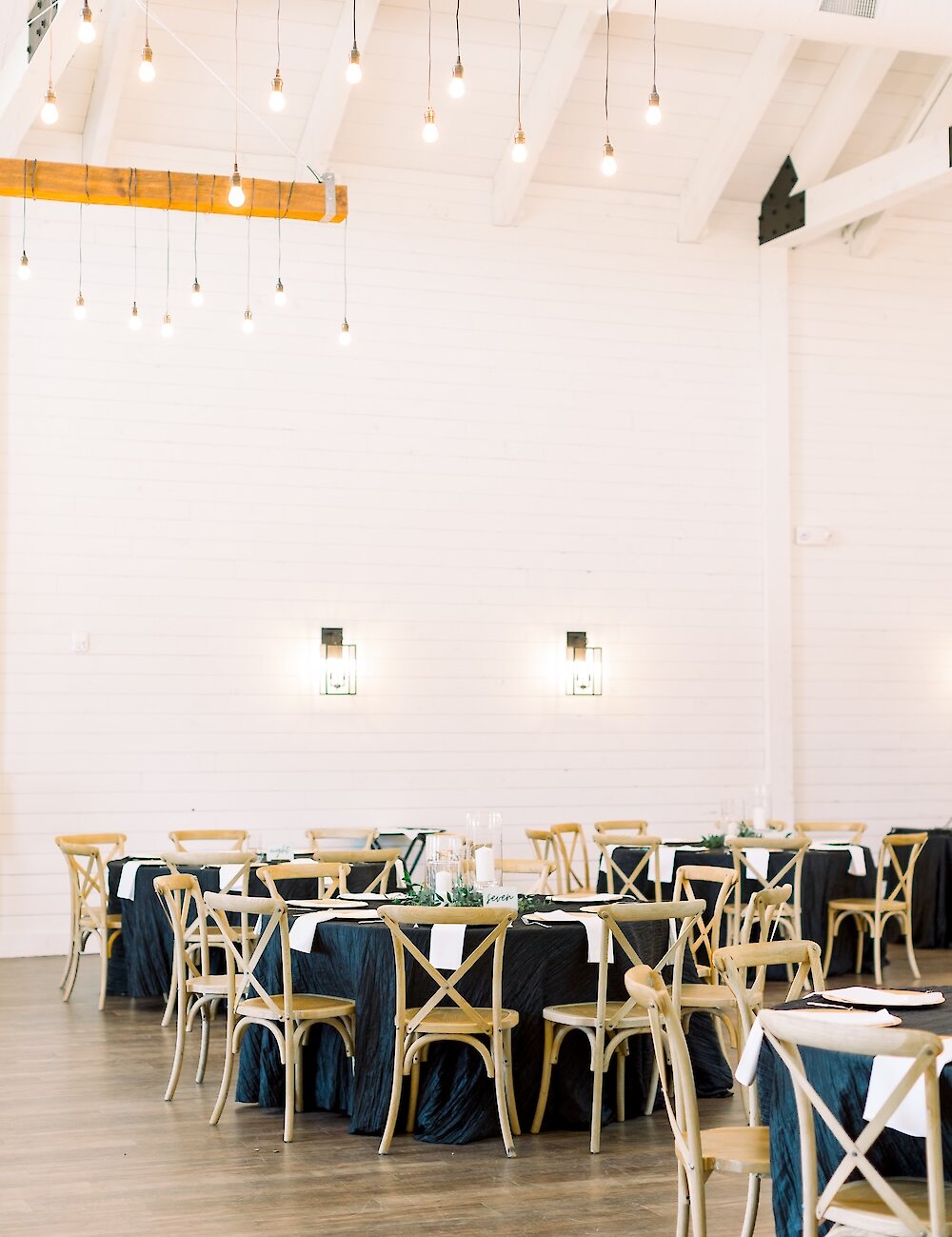 A rustic modern reception hall with white shiplap walls, exposed wood beams, string lights, and tables covered with dark black linen and wooden cross-back chairs.