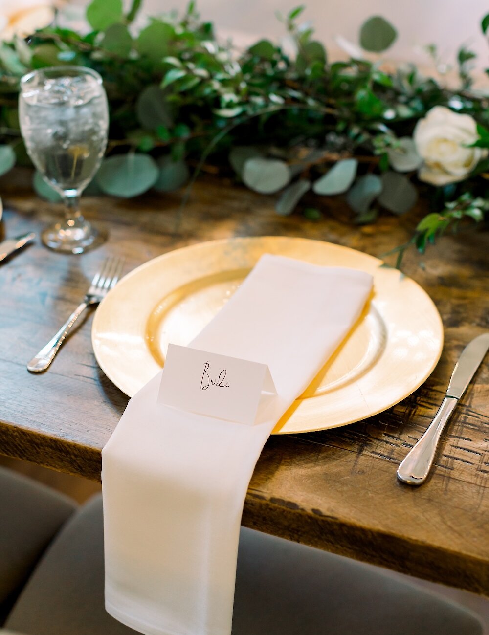 Close-up of a rustic wooden reception table place setting featuring a gold charger plate, white napkin, water glass, and a "Bride" place card with eucalyptus garland.