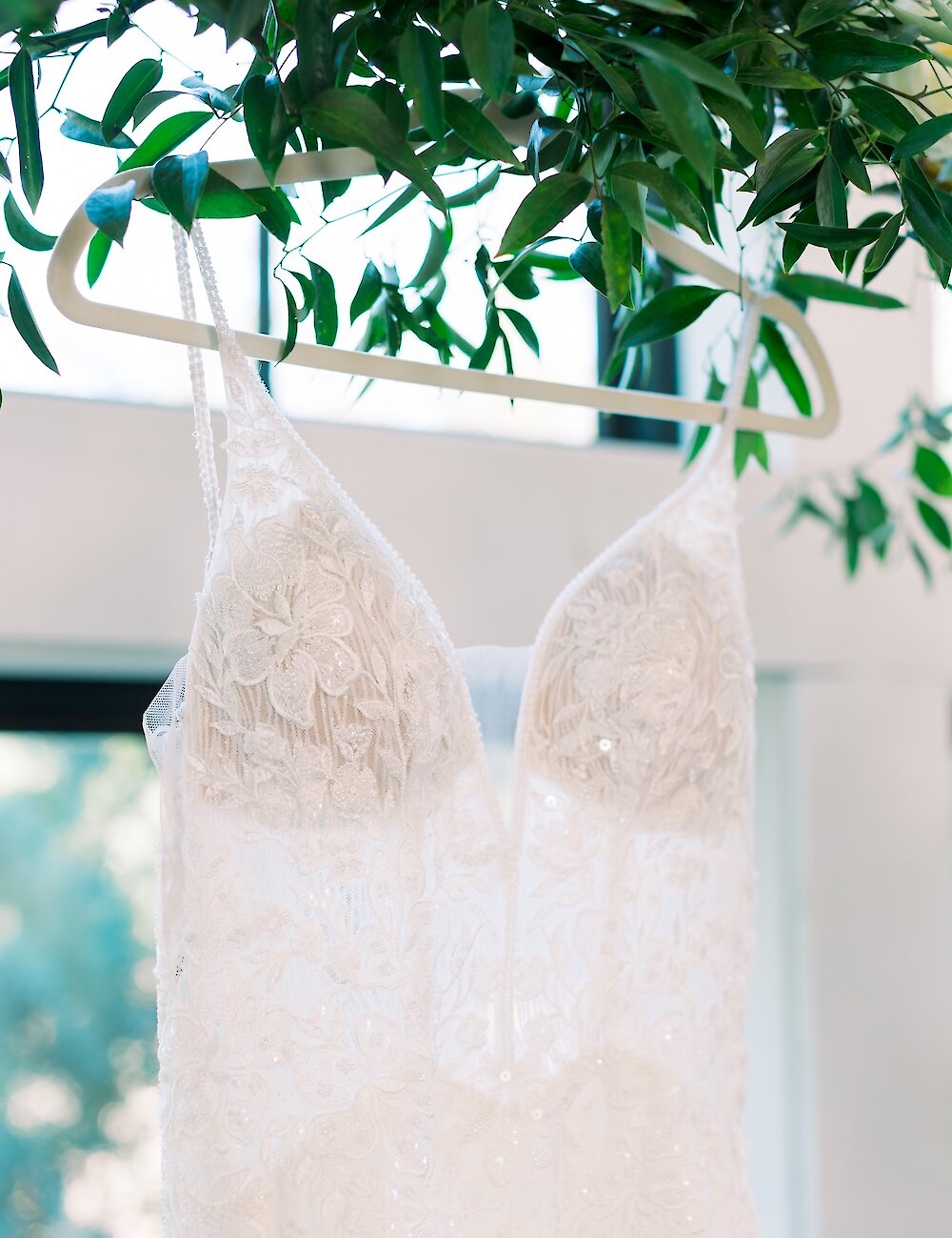 Close-up of a delicate white lace and beaded wedding dress with spaghetti straps, hanging on a hanger surrounded by lush green foliage.