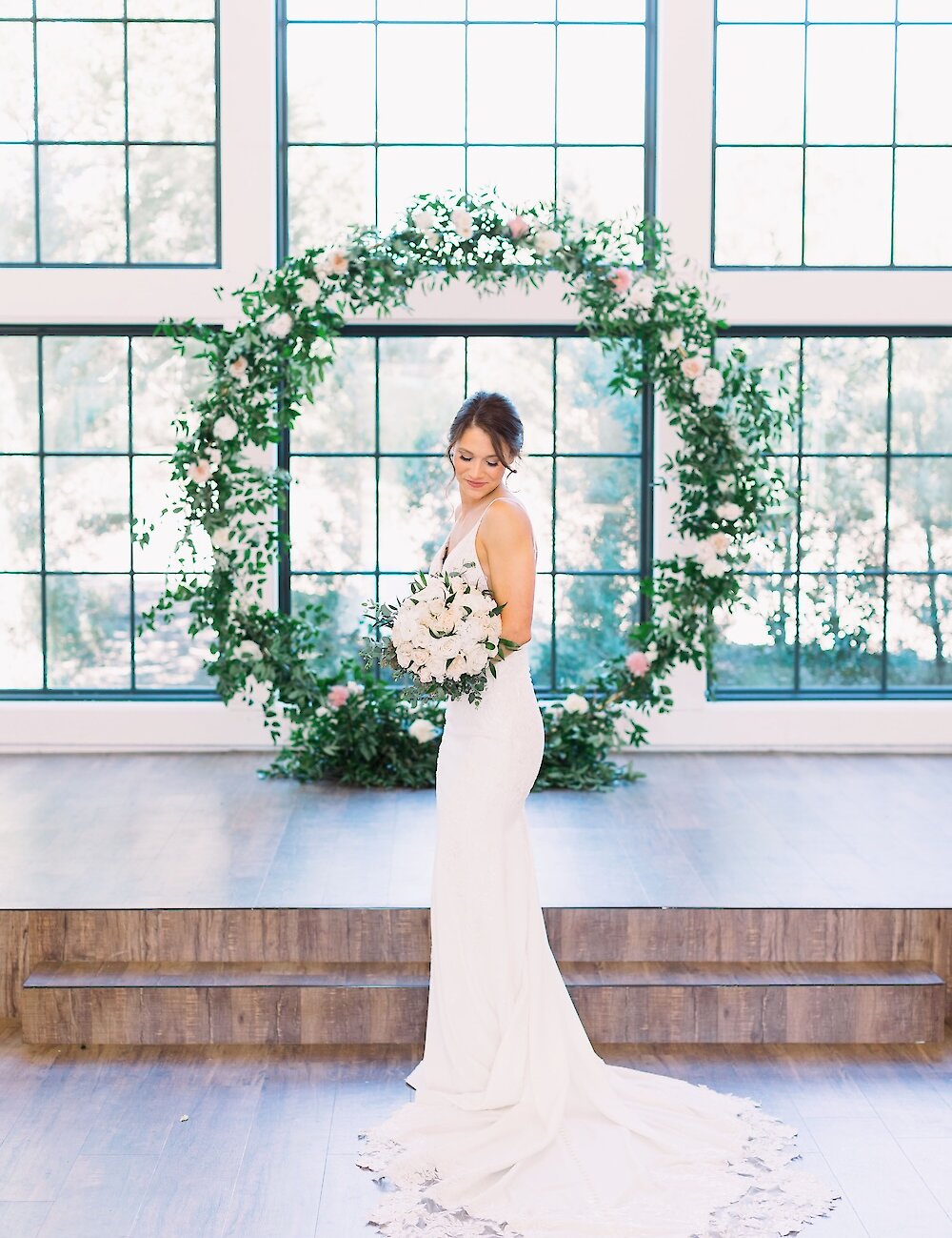 The bride in a fitted white gown with a lace-trimmed train poses, holding a bouquet, in front of a circular floral arch and large grid windows.