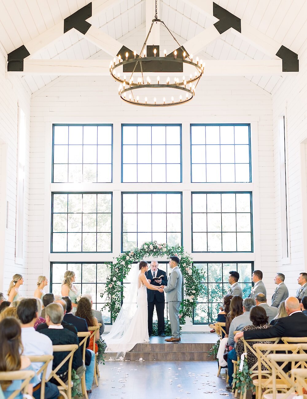 The bride and groom exchange vows in a white rustic chapel with vaulted ceilings, large grid windows, and a circular green and white floral altar.