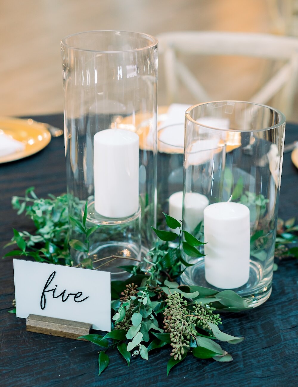 A close-up of a reception table centerpiece featuring a 'five' table number card next to white candles encased in clear hurricane vases and surrounded by eucalyptus and greenery.