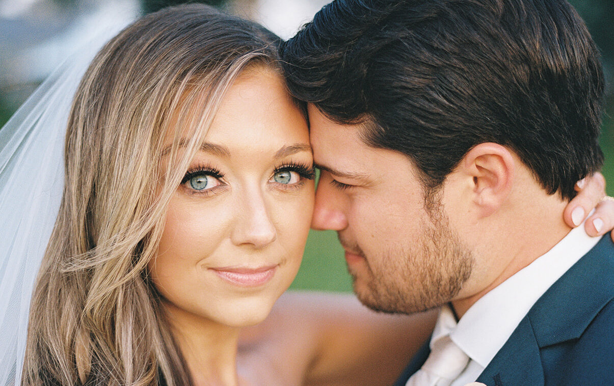 A close-up portrait of the beautiful bride with bright blue eyes looking at the camera while the groom, in a navy suit, leans his head against hers.