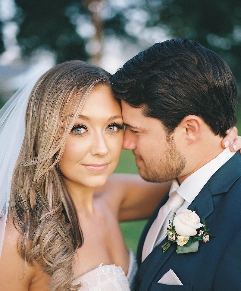A close-up portrait of the beautiful bride with bright blue eyes looking at the camera while the groom, in a navy suit, leans his head against hers.
