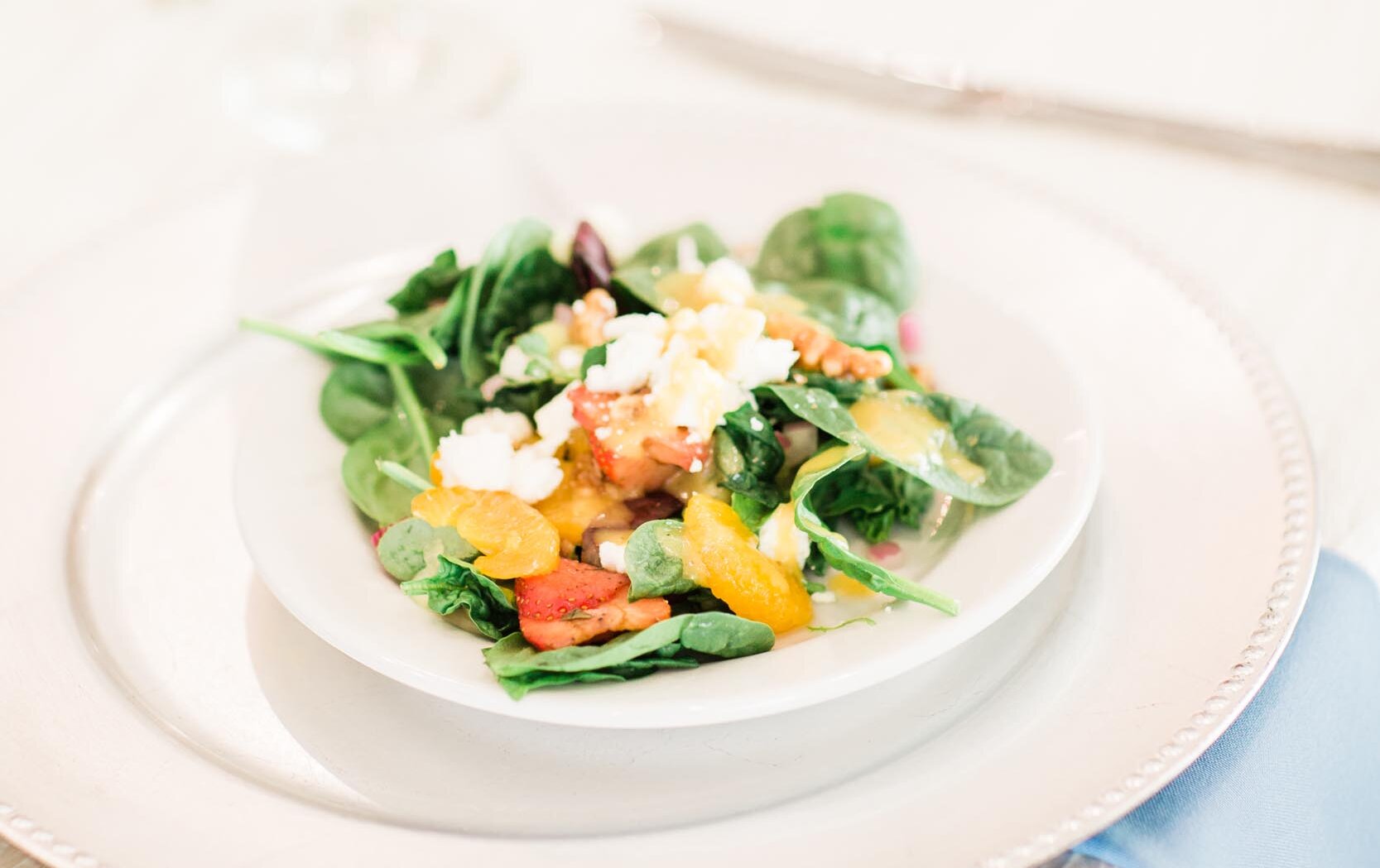 A freshly plated wedding salad starter featuring spinach, feta cheese, fruit, and citrus on a white plate with a decorative charger and a light blue napkin.