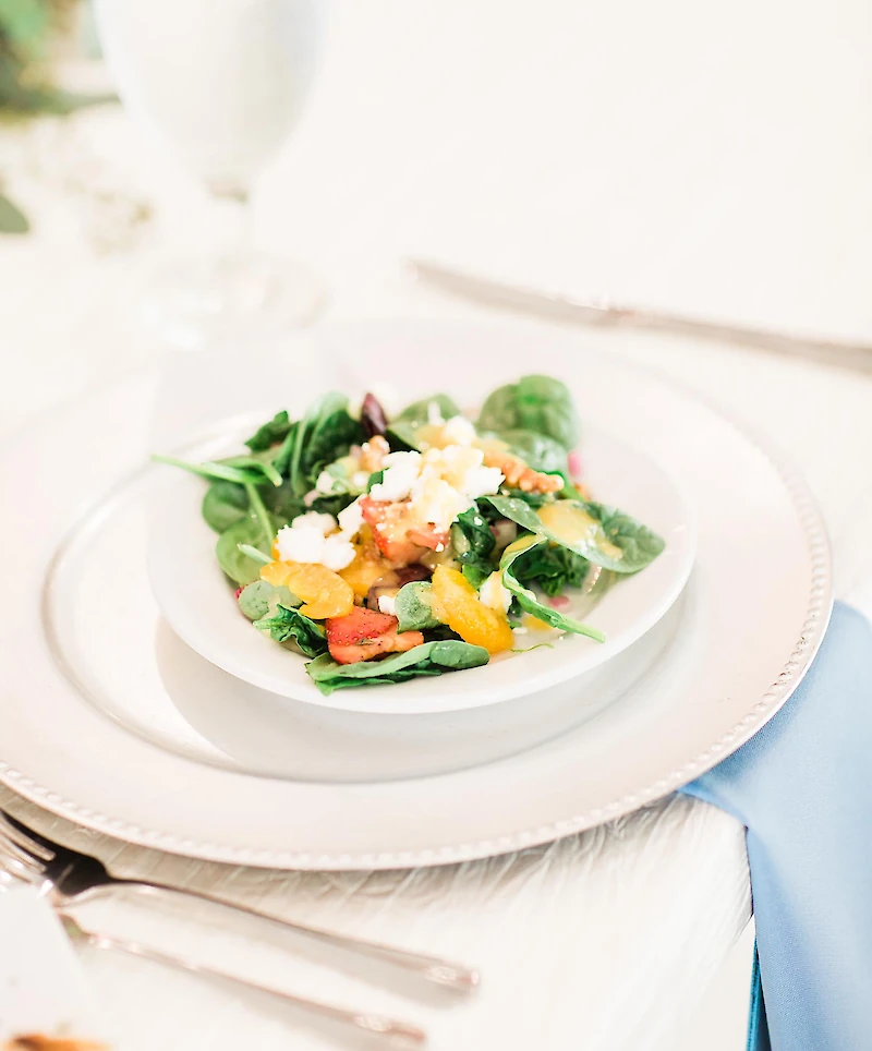 A freshly plated wedding salad starter featuring spinach, feta cheese, fruit, and citrus on a white plate with a decorative charger and a light blue napkin.