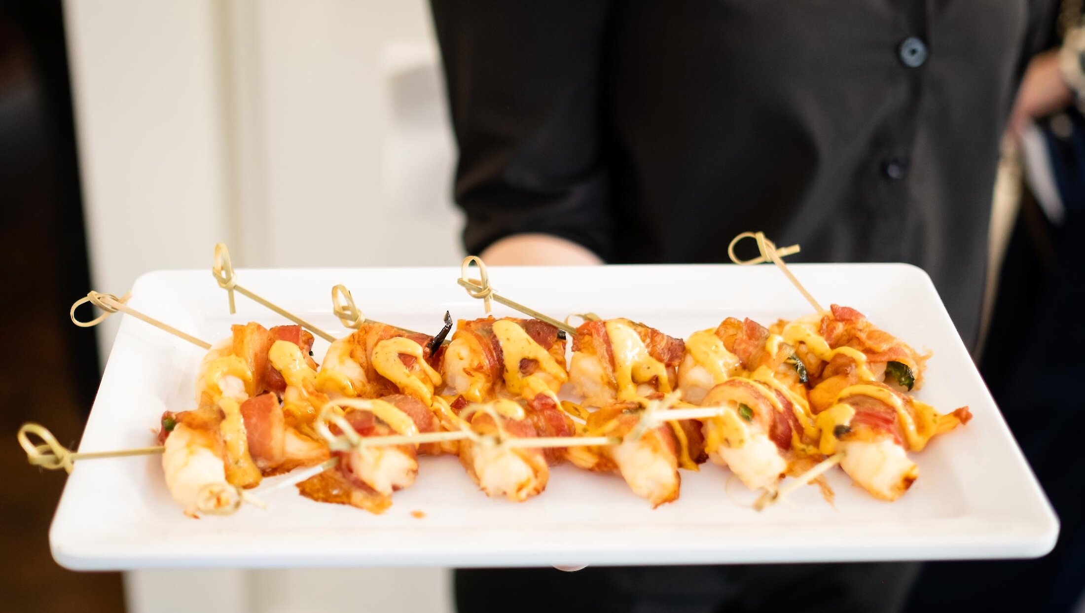 A server holds a white platter of catering appetizers consisting of bacon-wrapped shrimp skewers drizzled with a mustard sauce.