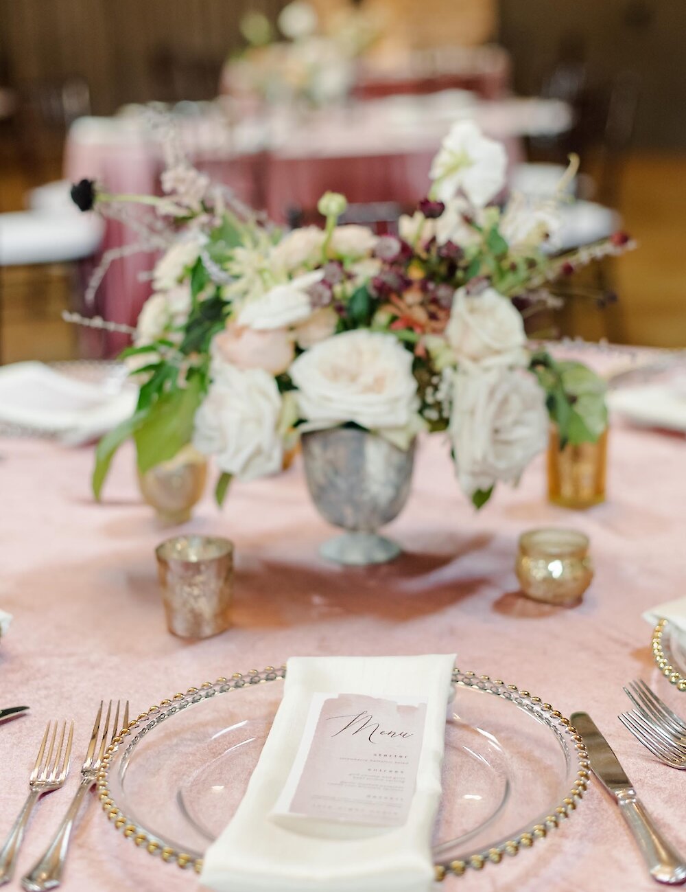 A wedding table setting featuring a beaded glass charger plate, a white menu card, and a floral centerpiece on a textured blush pink velvet tablecloth.