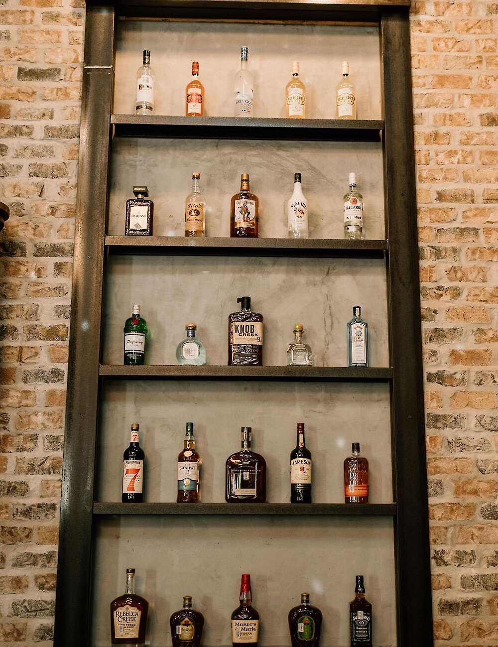 A tall, industrial metal shelving unit is mounted on a brick wall, displaying rows of various liquor and spirit bottles at the wedding reception bar.