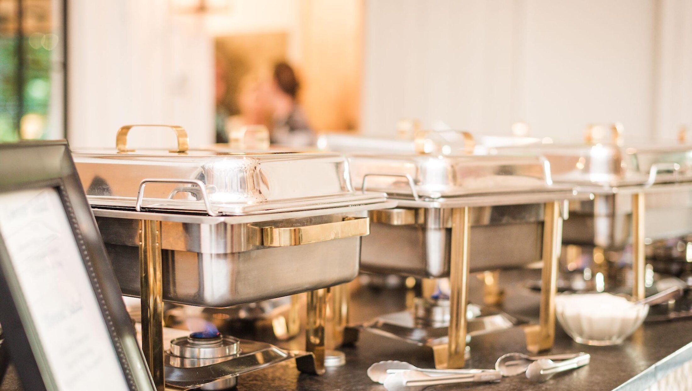 A close-up view of three professional stainless steel chafing dishes with gold accents set up on a long dark counter for the wedding buffet line.