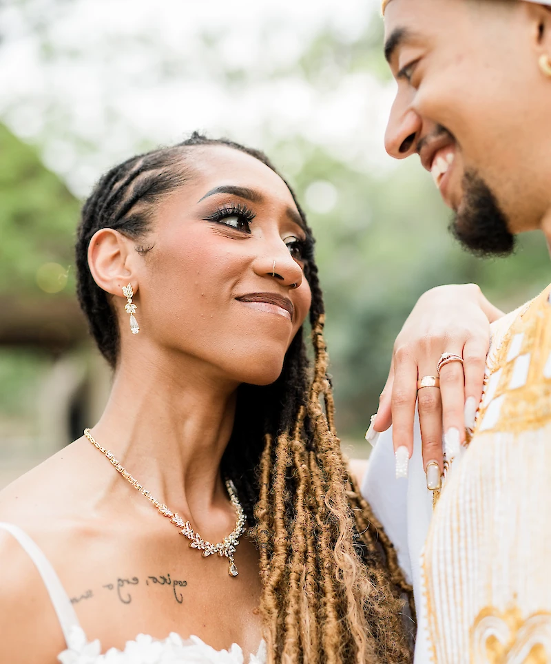 Close-up of the beautiful bride looking up at the groom, showcasing her necklace, earrings, and the rings on his hand, which is resting on her shoulder.