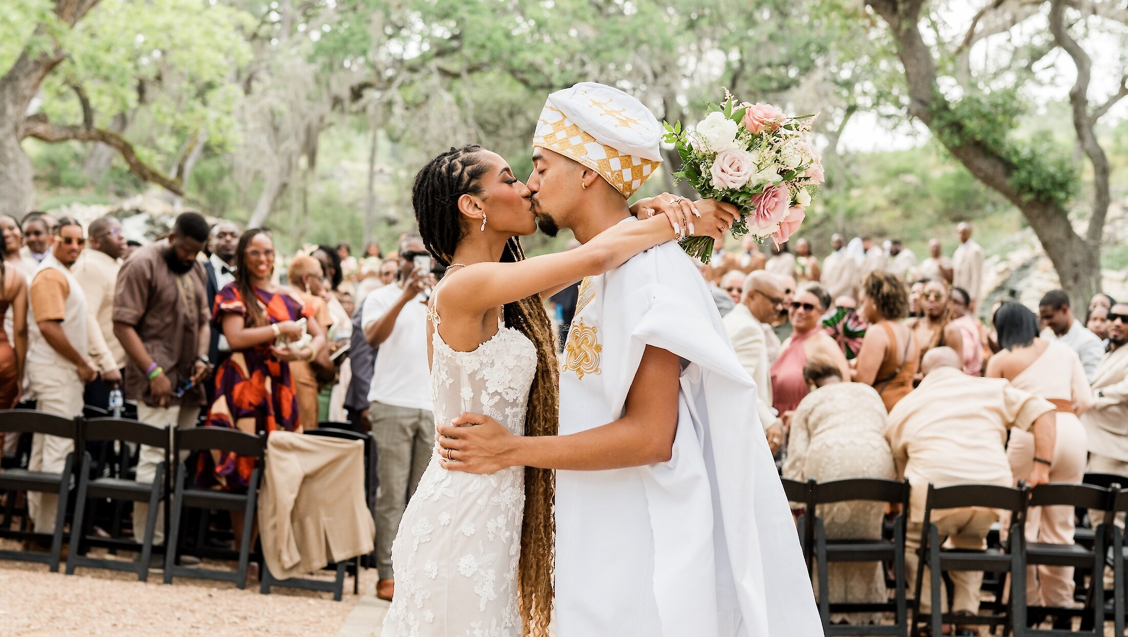 The bride and groom share a passionate kiss after the ceremony, with the groom in white and gold traditional attire and the bride in a lace gown, holding a pink and white bouquet.