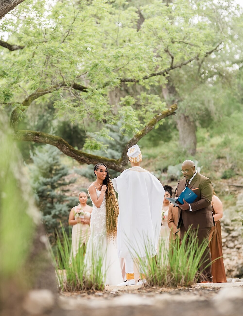 The bride looks at the groom during their outdoor ceremony, with the groom wearing white traditional attire and the officiant in a brown suit nearby.