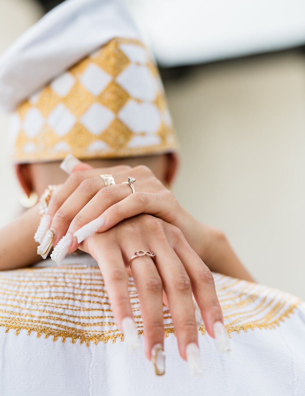 Close-up of the bride's hands with long manicured nails resting on the groom's shoulder, showcasing her rings and bracelets against his white and gold embroidered traditional attire.