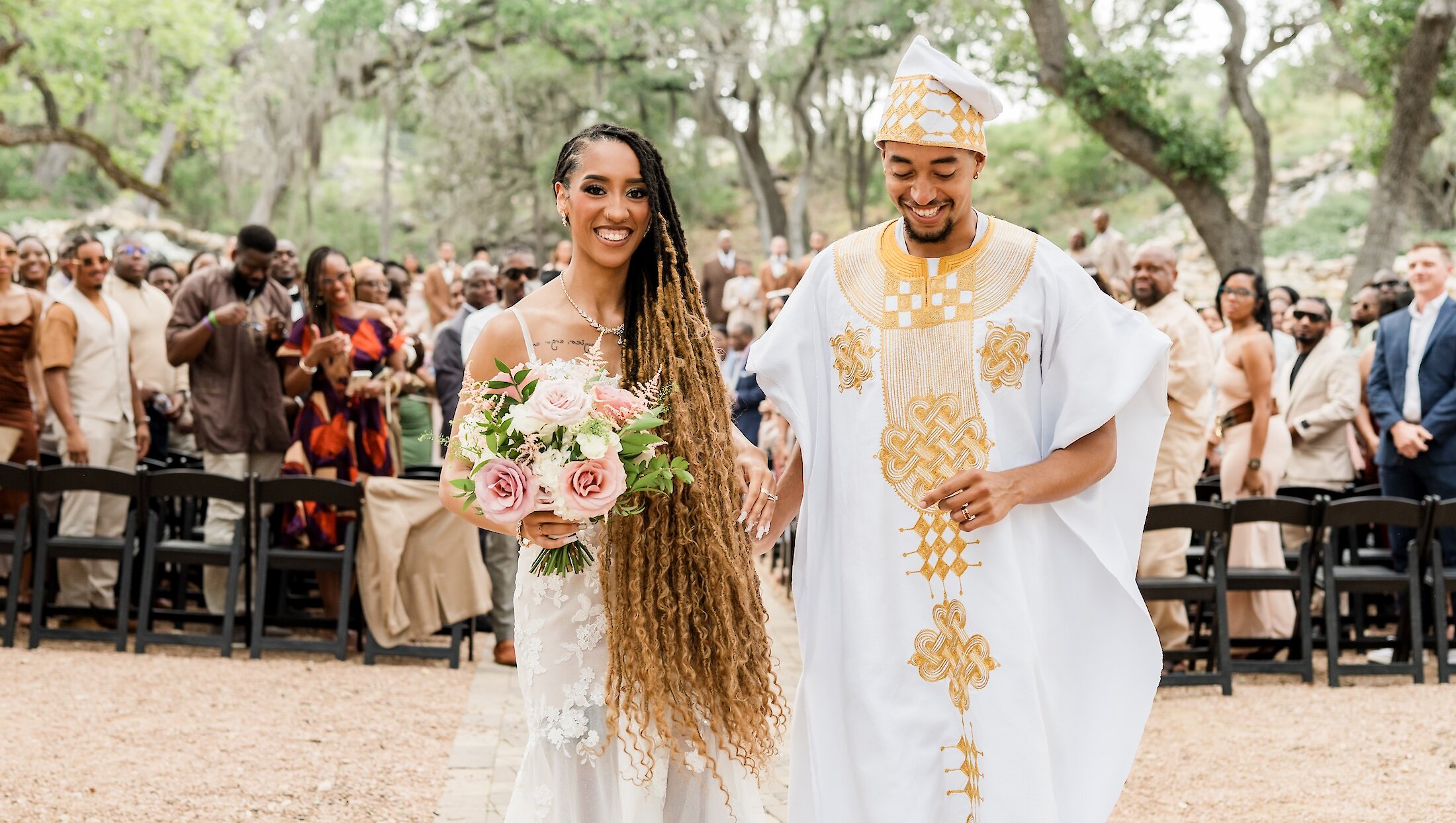 The smiling bride and groom walk down the outdoor aisle, with the groom wearing white and gold African traditional attire and the bride holding a large pink and white bouquet.